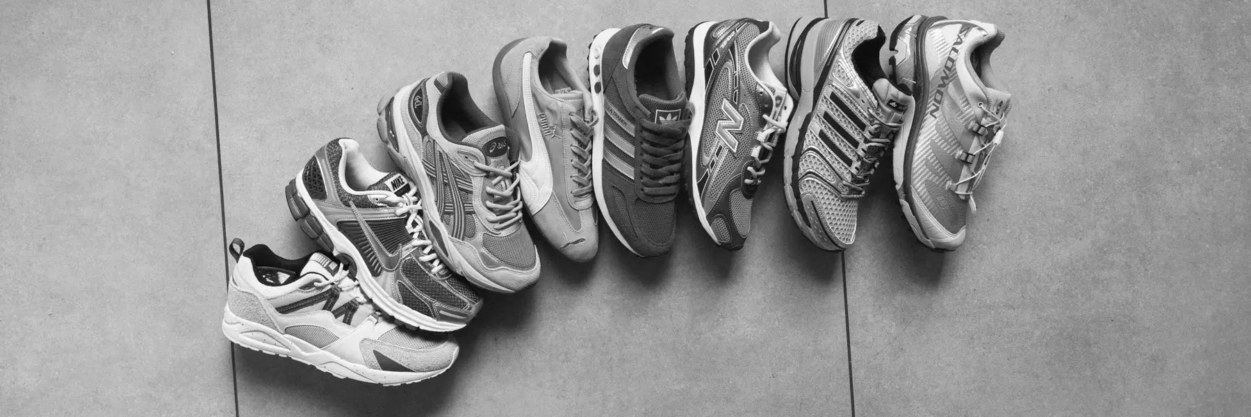A row of eight different athletic shoes is neatly arranged on a tiled floor, viewed from above. The image is in black and white, conveying a classic feel. Desktop Banner