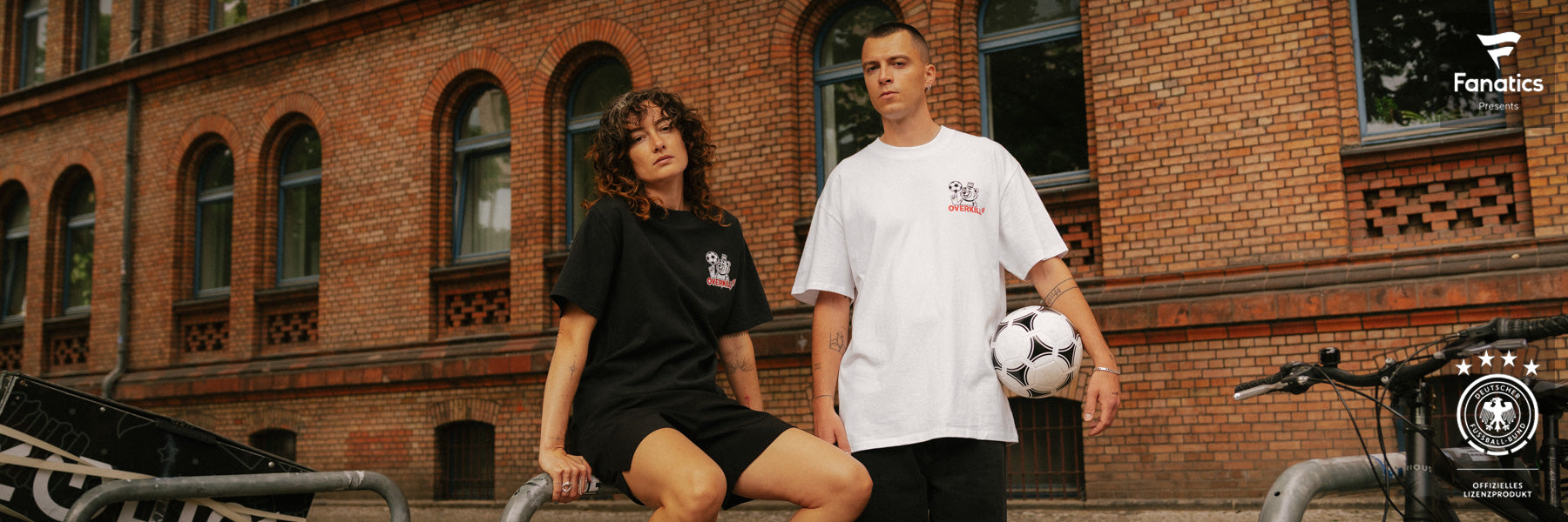Two people in casual sportswear pose confidently outside a brick building. One sits on a railing, the other stands holding a soccer ball. Both wear casual streetwear styles amidst an urban backdrop.