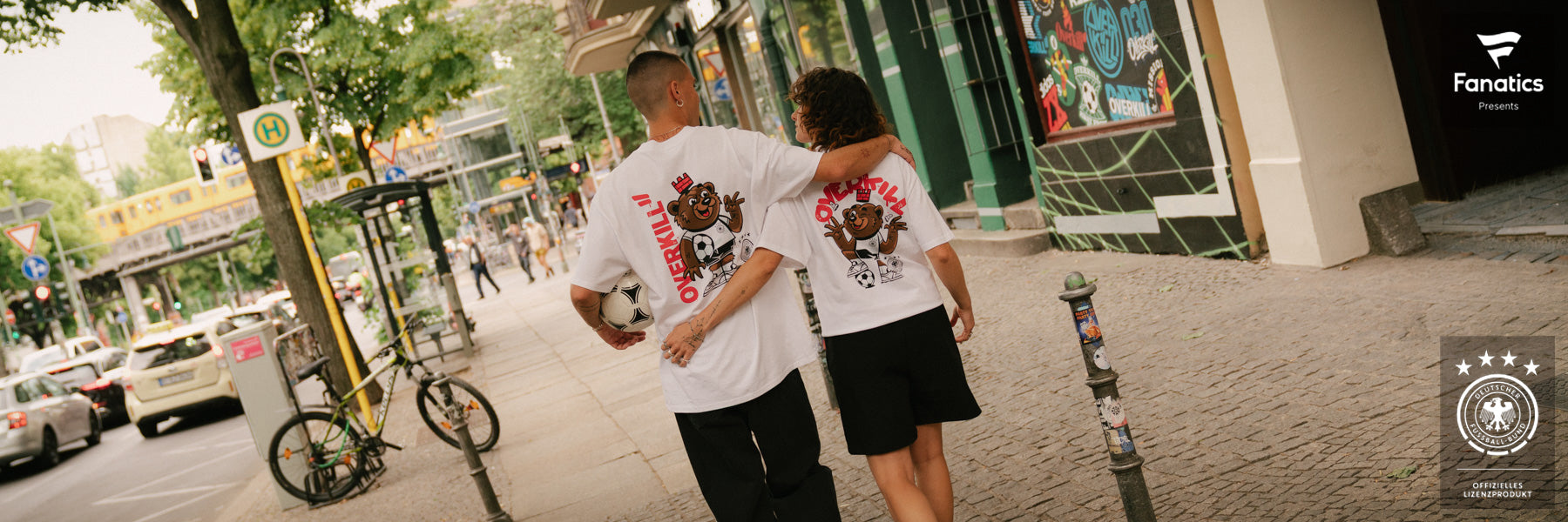 A couple walks down a city street, wearing matching white "OVERKILL x DFB" t-shirts with a cartoon bear and soccer motifs. They hold a soccer ball, exuding a relaxed, sporty vibe.