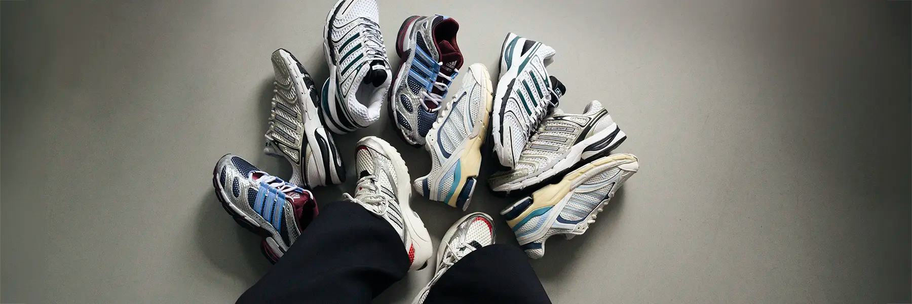 A collection of assorted adidas Y2k running shoes surrounds a pair of black pants-clad legs on a gray background. The sneakers vary in color, with a predominantly blue and white theme, conveying a sense of choice and style exploration.