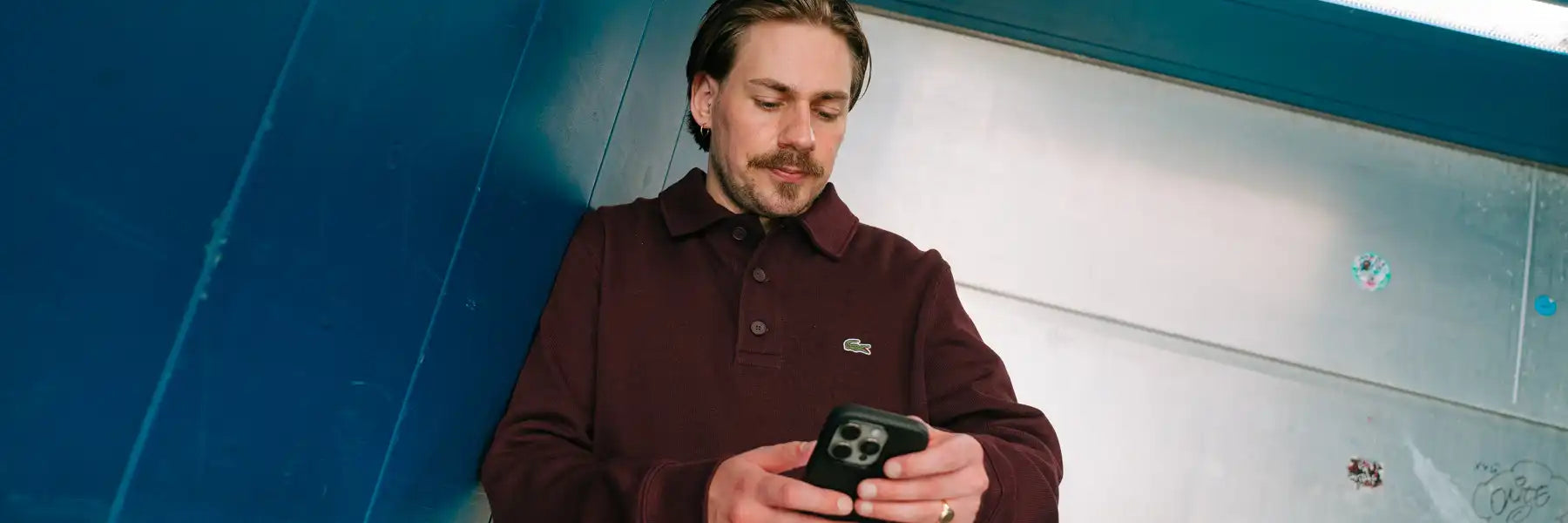 Man with mustache and earrings wearing a dark polo shirt with a Lacoste logo looking at a smartphone