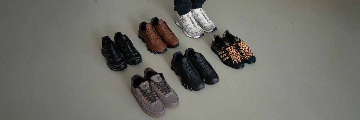 Six pairs of autumn sneakers arranged on the floor with a person wearing white sneakers standing behind them