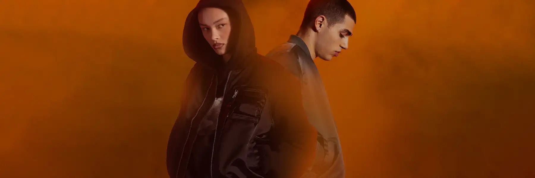 Two individuals stand back-to-back against an orange smoky background, one in a hooded jacket looking forward and the other in a gray jacket looking down. Moody atmosphere. Both wearing Alpha Industries UV jackets