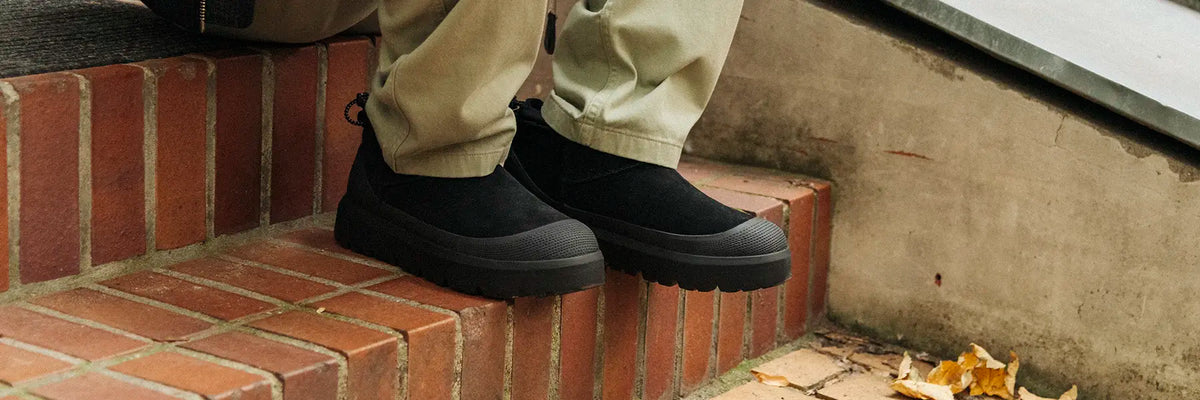 Person wearing black slip-on boots  from UGG with thick soles sitting on brick steps