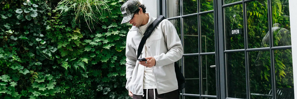 Man wearing a light fleece jacket, camouflage cap, and crossbody bag looking at his phone outdoors near a window and green foliage