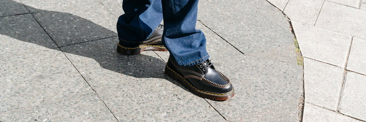 Person wearing black leather shoes with yellow stitching from Dr. Martens and blue jeans standing on a tiled pavement