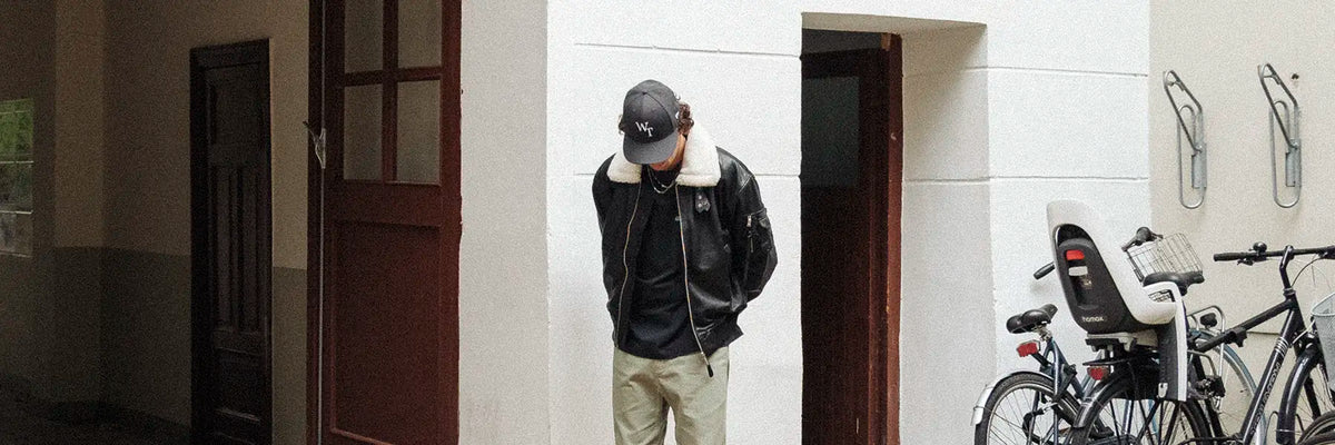 Person wearing a black leather jacket with a white shearling collar, black cap with 'WT' letters, black shirt, and light-colored pants, standing with head down
