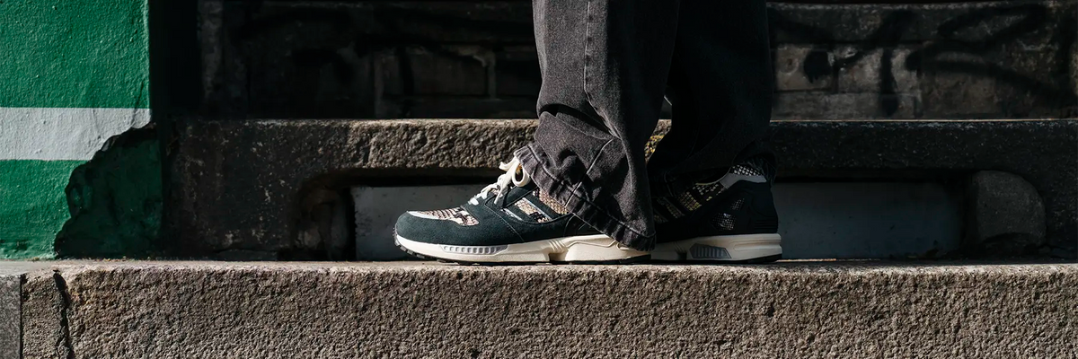 A close-up lifestyle shot of a person's legs and feet wearing a pair of ZX 8000 adidas sneakers in animal print, standing on concrete steps.