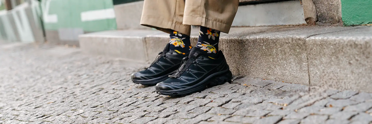 Person wearing black Salomon sneakers with patterned socks and beige pants standing on cobblestone steps