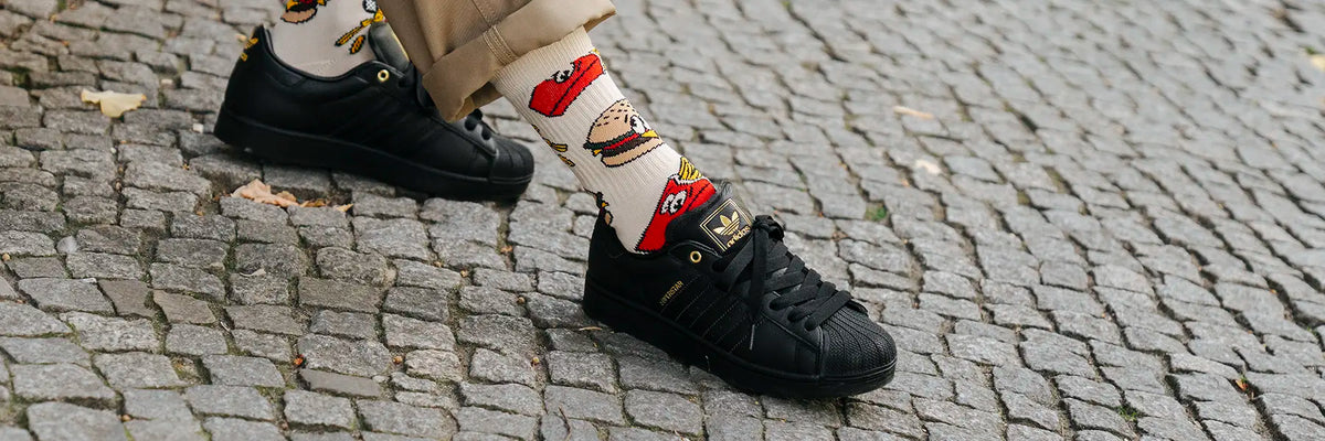 Foot wearing black Adidas Superstar sneaker and white Stance sock with cartoon fries and burger pattern on cobblestone pavement