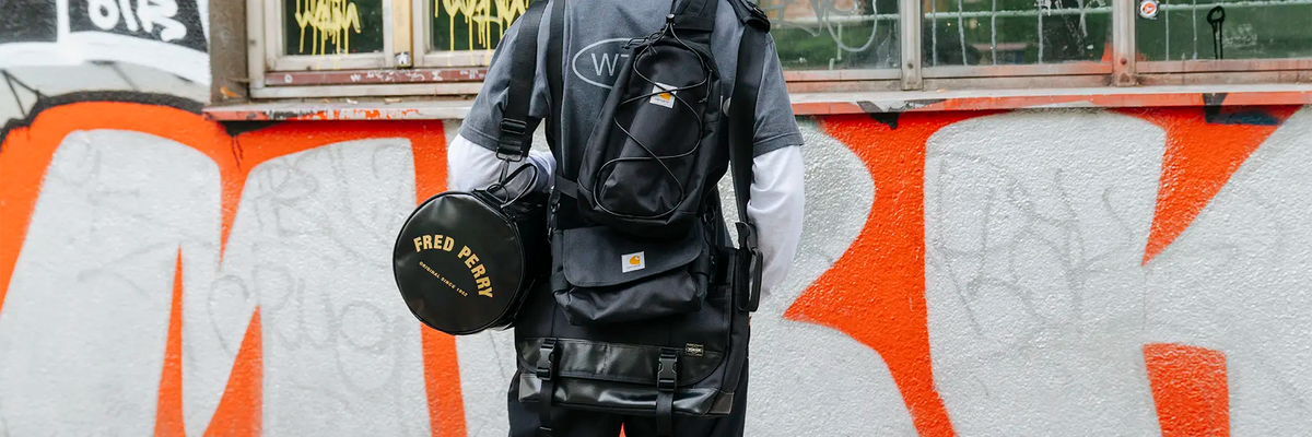 Person seen from behind wearing a dark short-sleeve shirt over a white long-sleeve shirt carrying multiple black bags & backpacks including a round bag with 'FRED PERRY' text and a rectangular backpack with a Carhartt logo patch