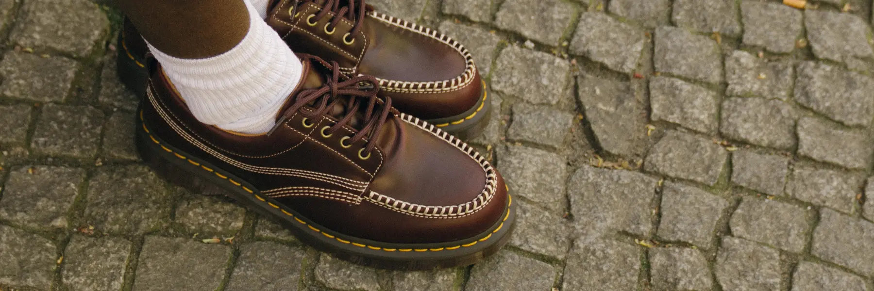 Brown leather Dr. Martens shoes with white stitching and yellow threading are worn with white socks on a cobblestone street, conveying a casual, stylish vibe.