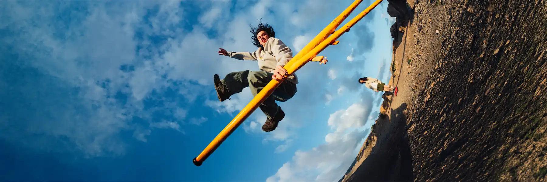 A person joyfully jumps with two long yellow poles in an expansive landscape. A bright blue sky with fluffy clouds enhances the dynamic scene. Banner for Gramicci