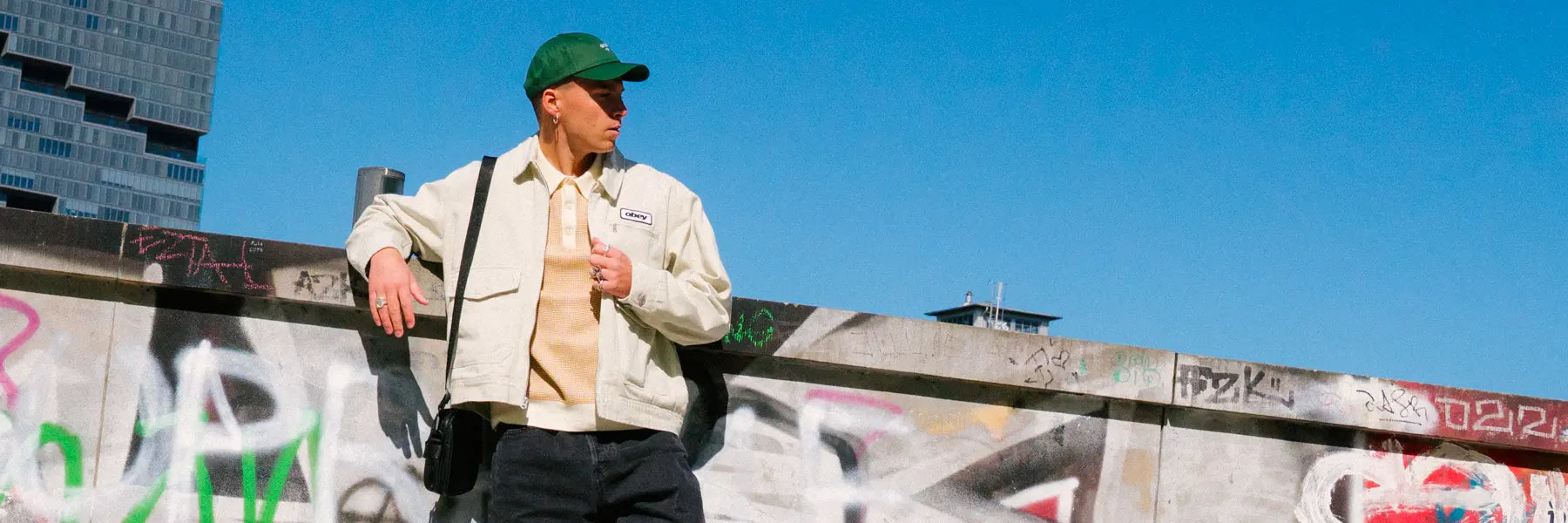 A person in a green cap and casual Obey jacket leans against a graffitied wall, set against a clear blue sky and urban buildings, conveying a relaxed, urban vibe.