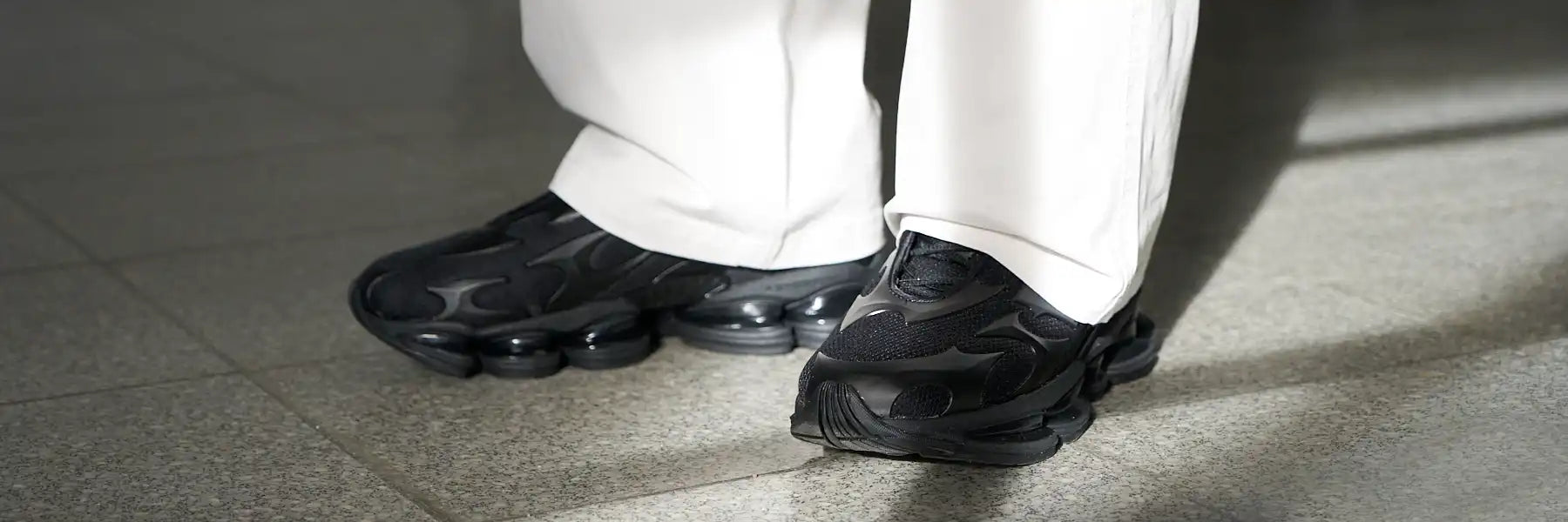 Person wearing black New Balance 2000 sneakers with bold patterns and thick soles, standing on a tiled floor. The light hits the shoes, creating a stylish and modern vibe.
