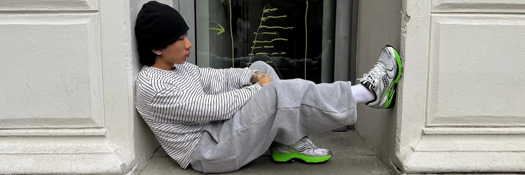 Person in gray sweatpants, striped sweater, and black beanie sitting on the ground with feet resting against a door frame wearing gray and green Saucony running shoes