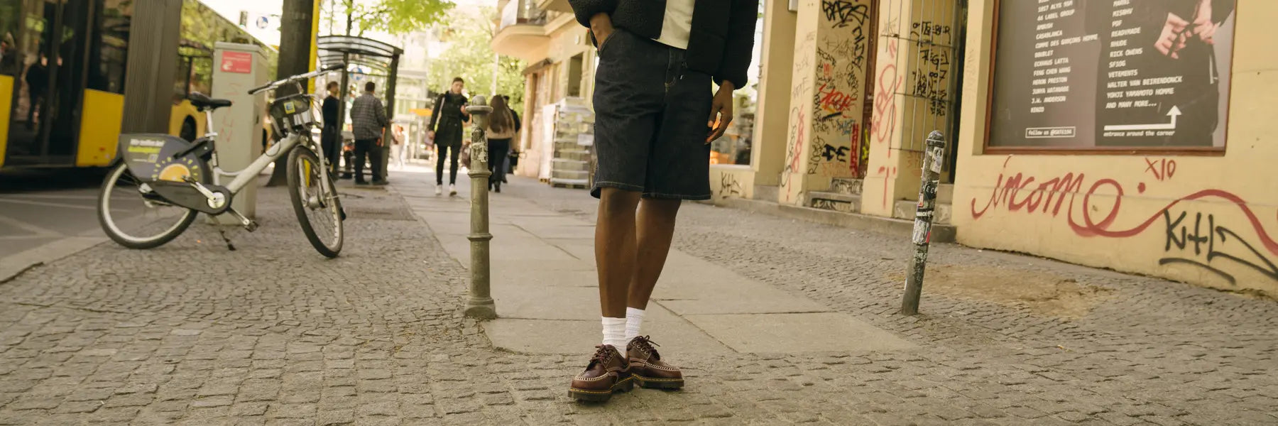 A person stands on a graffiti-covered urban street, wearing a black jacket, denim shorts, white socks, and brown shoes. The mood is casual and relaxed. Desktop banner