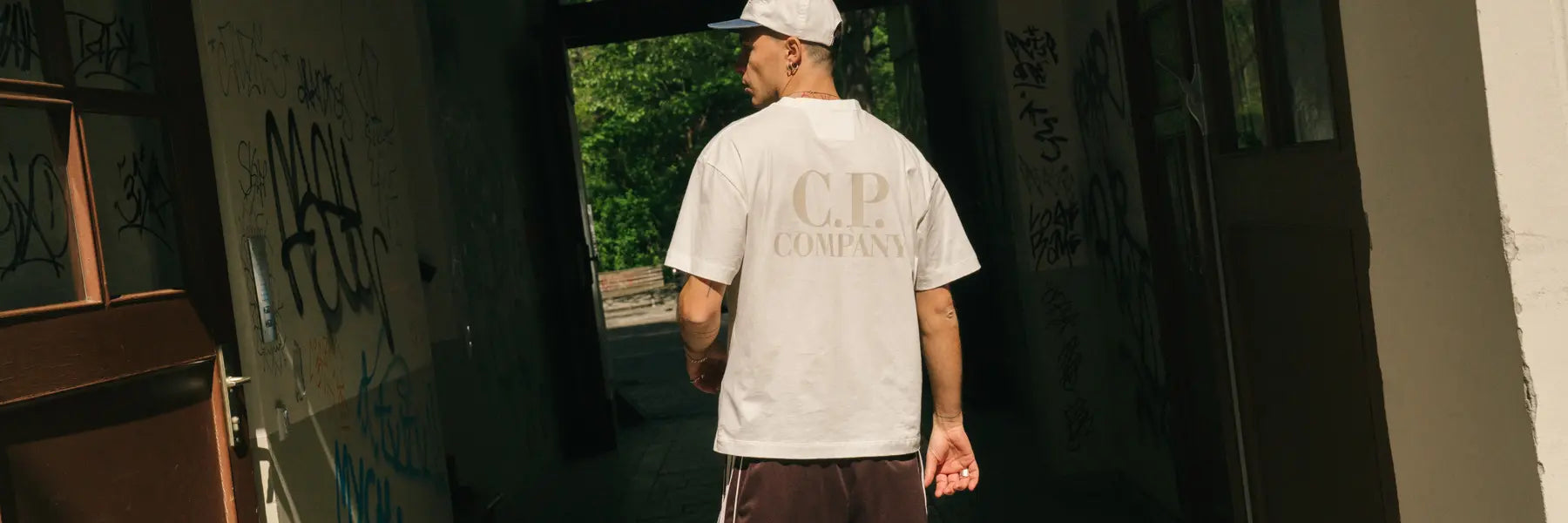 A man in a white "C.P. Company" T-shirt and cap walks through a graffiti-covered corridor leading to a sunlit path surrounded by greenery.