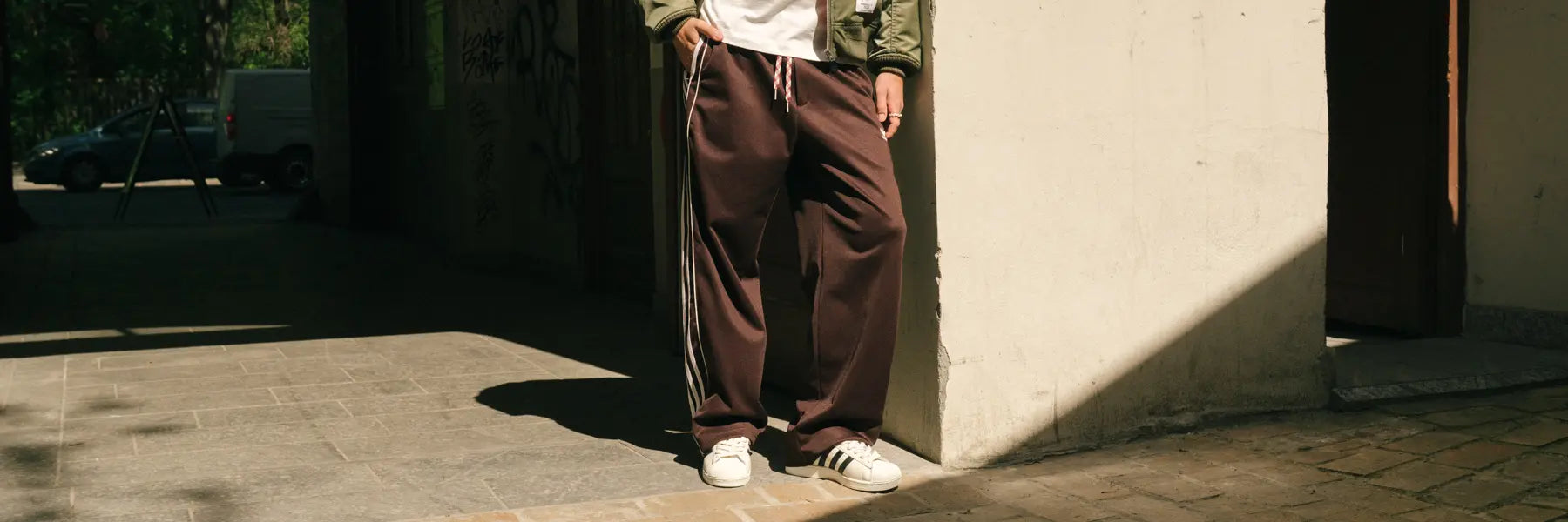 Person stands at the corner of a sunlit building, wearing brown track pants, a jacket, and white sneakers; shadows cast a relaxed, casual tone. Desktop Banner