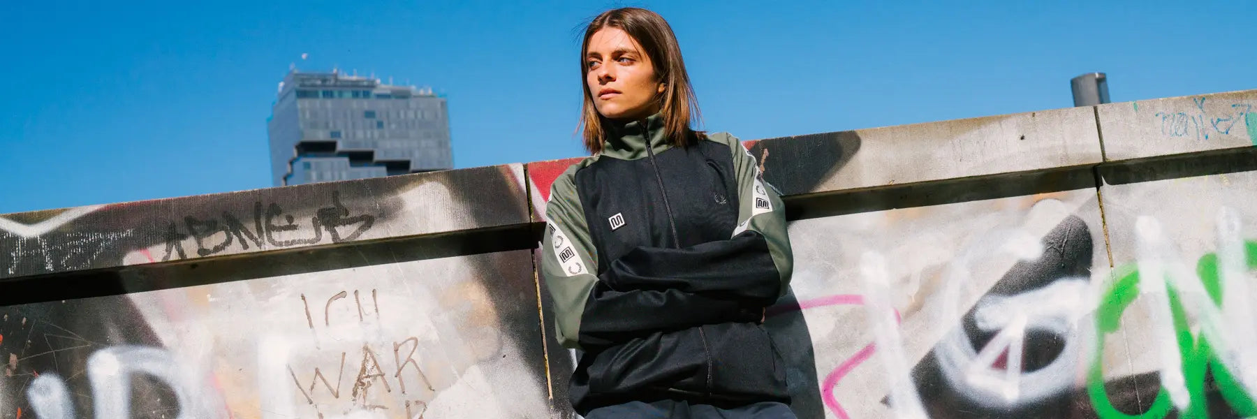 Young woman in a black and green jacket leans against a graffiti-covered wall, looking contemplative. Bright blue sky enhances the urban feel.