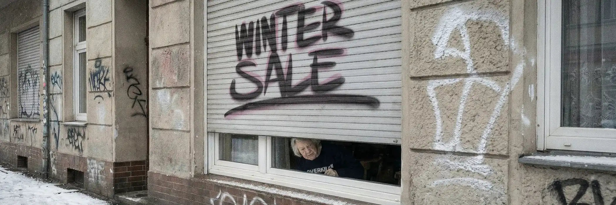 Closed shutters with "Winter Sale" in bold graffiti cover a graffiti-lined building wall. A person peeks from beneath, adding intrigue to the snowy urban scene.