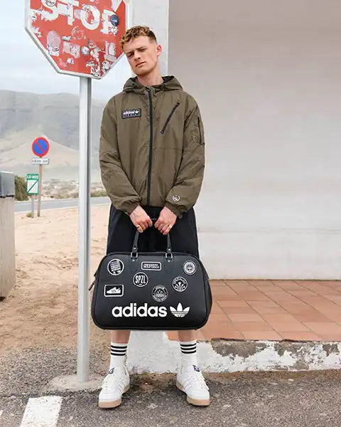 Young man in a green jacket and shorts stands against a weathered white wall, holding a black Adidas bag with various patches, next to a worn stop sign.