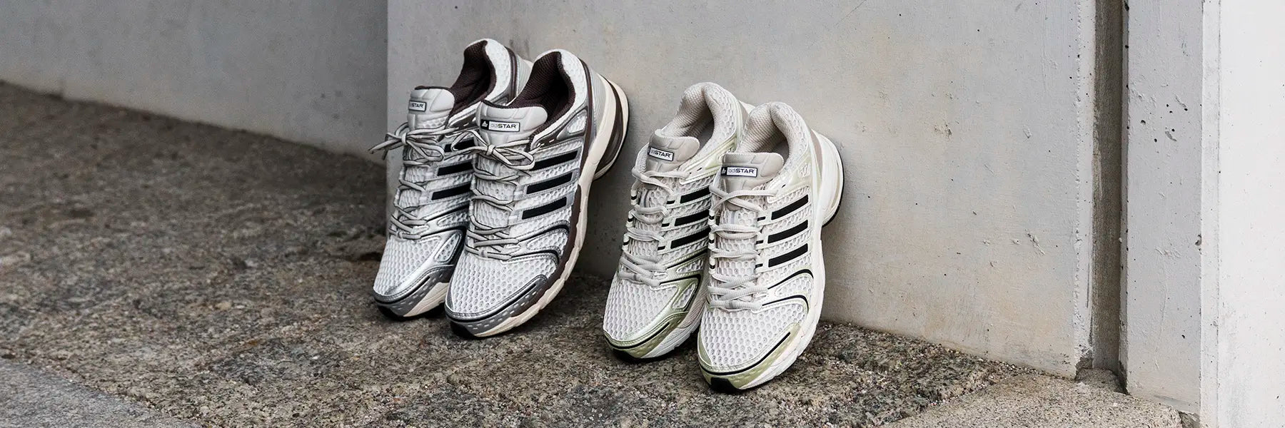 Two pairs of white adidas Adistar Control 5 sneakers with black stripes are placed against a textured concrete wall. The setting is outdoors, conveying a casual, relaxed vibe.