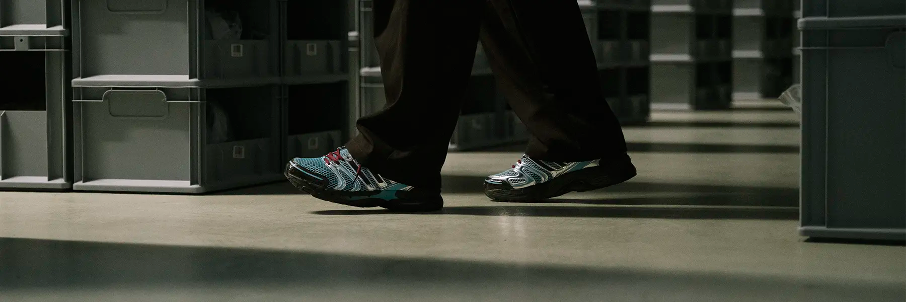 A person walks between rows of shelves filled with grey storage bins wearing adidas Adistar Control 5 Y2K Sneaker. Their colorful sneakers stand out against the neutral, industrial setting. Desktop Banner