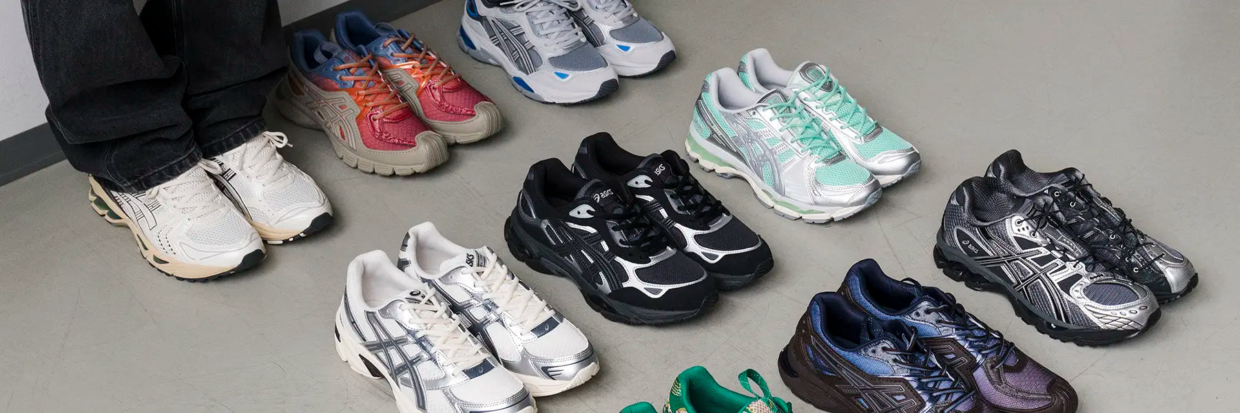 A collection of nine pairs of colorful sneakers, including white, black, red, and teal, arranged on a gray floor. Legs wearing white sneakers are visible on the left. Desktop Banner