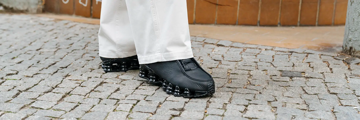 Person wearing black Nike Shox sneakers with segmented soles and white pants standing on cobblestone pavement