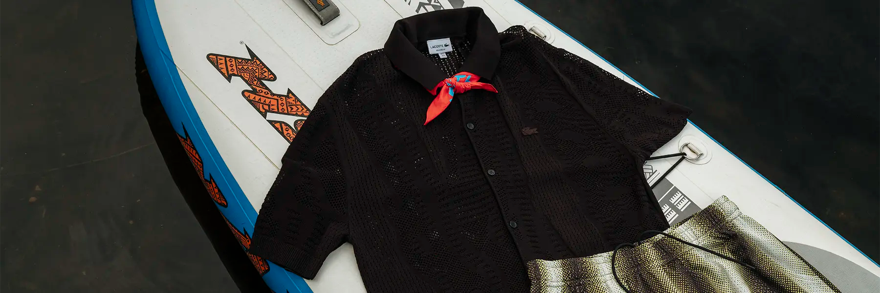 Black short-sleeved button-up shirt with a red bandana tied around the collar laid out on a surfboard next to metallic pleated shorts