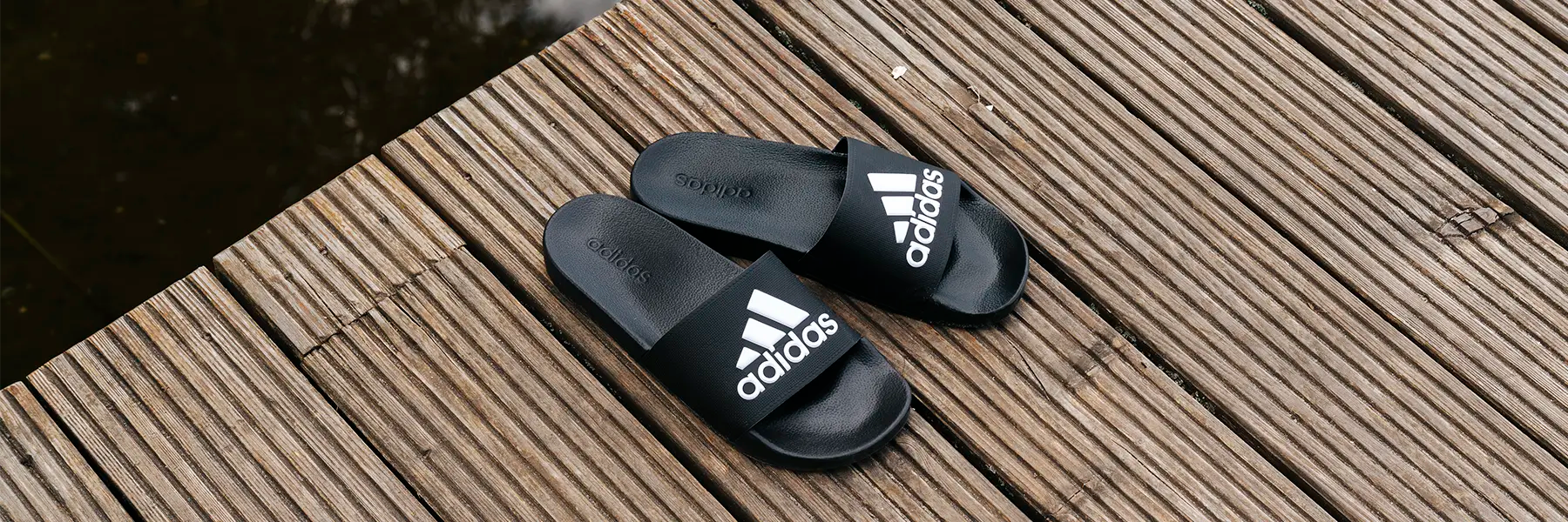 A pair of black Adidas slide sandals with white logos, placed on a wooden deck next to water.