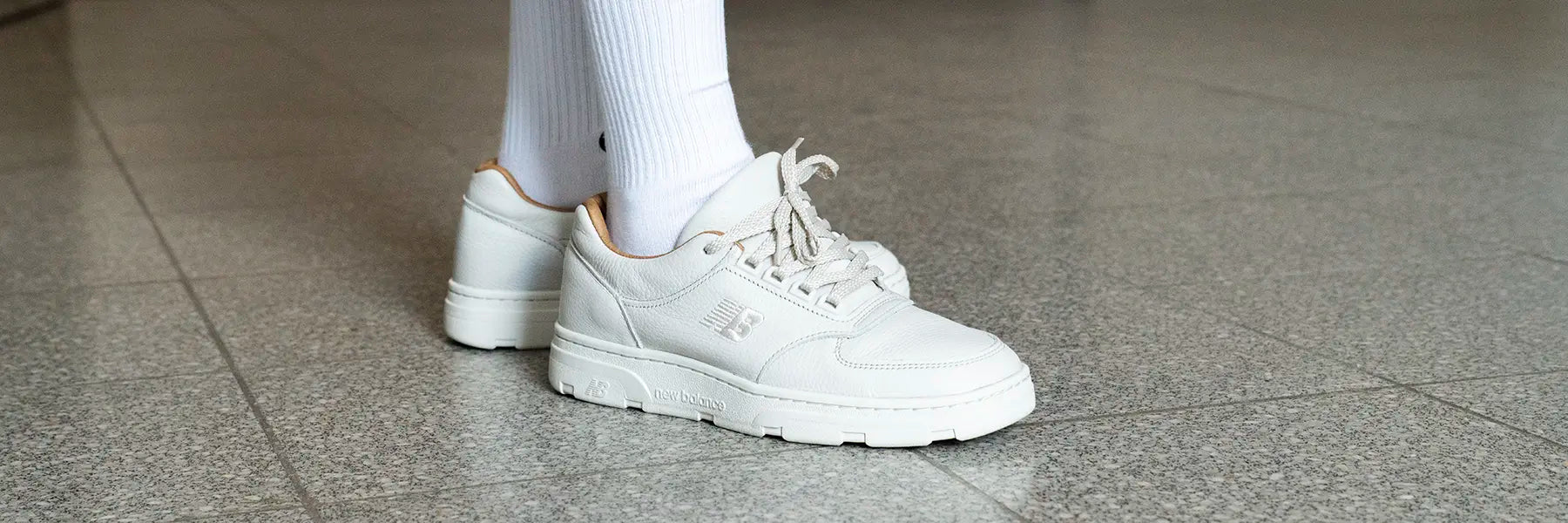 A person wearing white New Balance Allerdale sneakers and knee-high socks stands on a tiled floor. The image conveys a casual and minimalistic style.