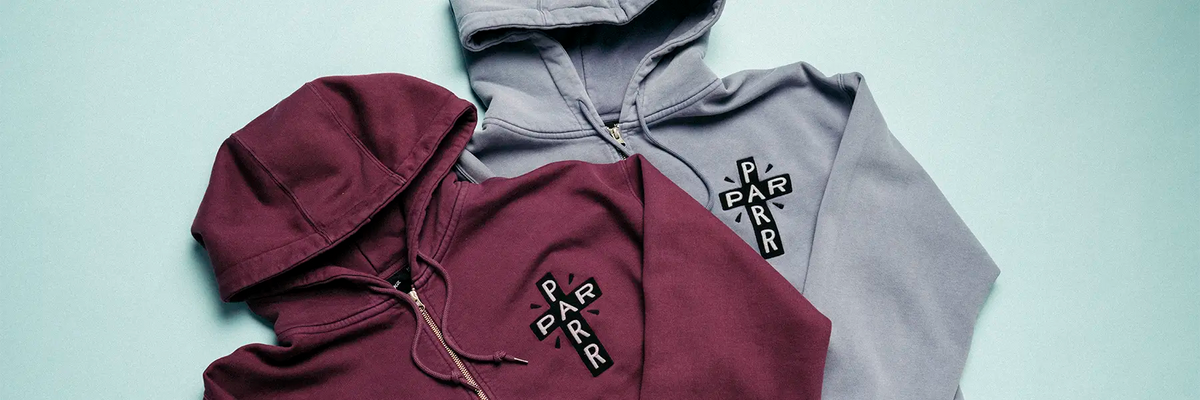 Two Parra hoodies arranged on a pale blue background: a burgundy hoodie in front with a black cross-shaped chest graphic and a gray hoodie behind it with a matching black cross graphic