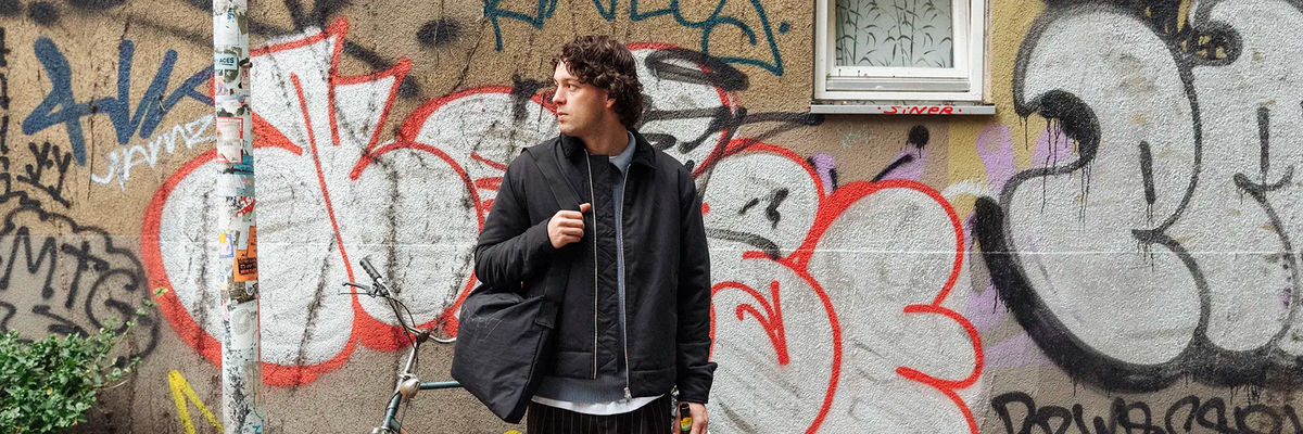 A man wearing a black zip-up jacket over a gray shirt stands in front of a graffiti-covered concrete wall.