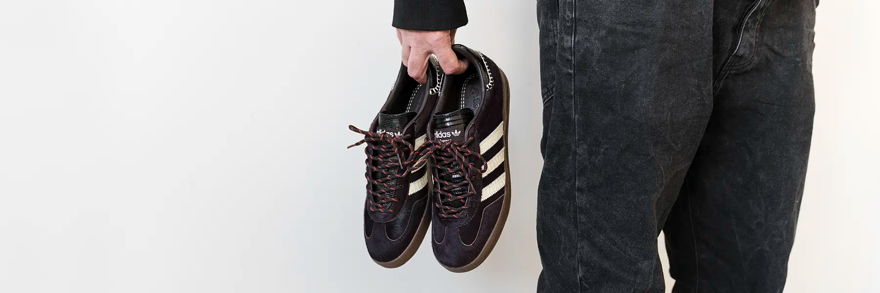 A person holds black and white Adidas sneakers by the laces in one hand. They are wearing dark jeans and a black sweater, standing against a plain white background.