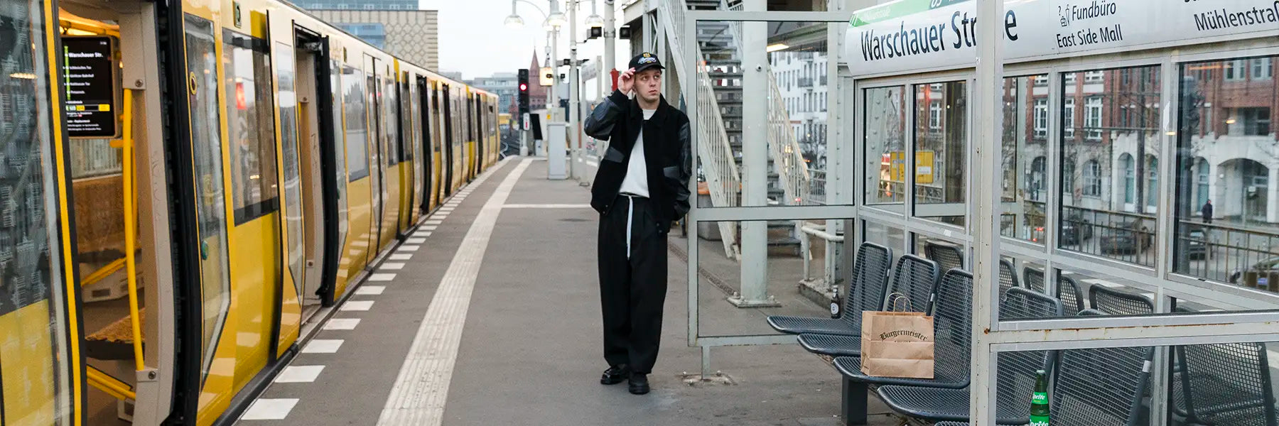 A person stands on the platform at a Berlin Train station in a black Outfit, beside a yellow train, with benches and a paper bag visible nearby.