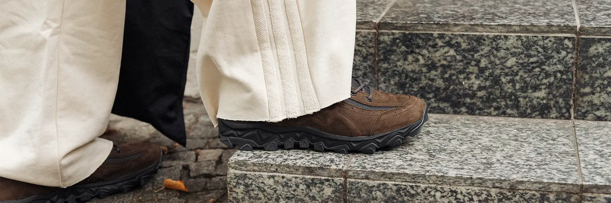 Brown suede sneaker with a textured black sole worn with cream-colored pleated pants stepping on stone stairs