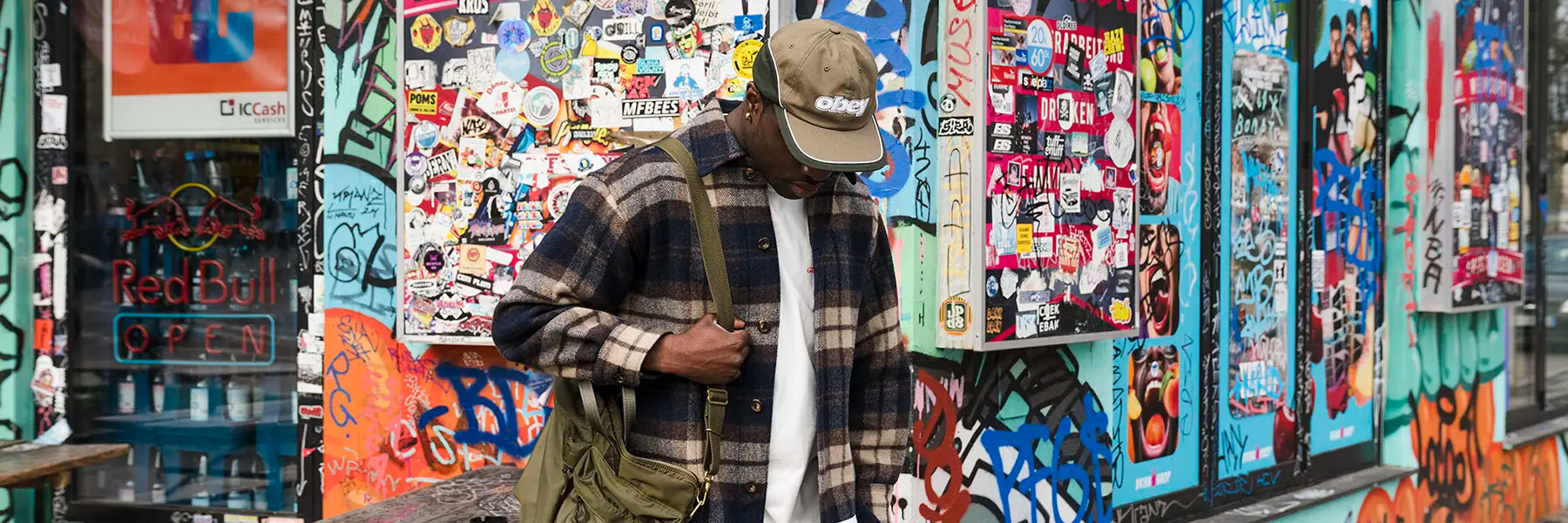 A person wearing a plaid jacket and olive cap stands in front of a vibrant, sticker-covered wall, holding a green bag. The scene feels urban and trendy.