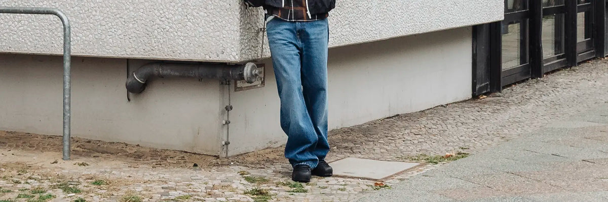 A person standing outdoors next to a textured concrete building corner, wearing blue jeans, a dark jacket, and dark shoes.