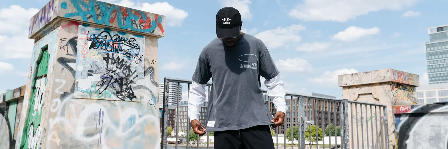 Man wearing a black cap and layered long-sleeve and short-sleeve shirts standing by a railing with urban buildings and water in the background