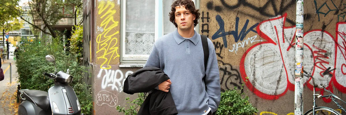 Man with curly hair wearing a blue long-sleeve polo shirt and striped pants holding a black jacket over his arm standing in front of a graffiti-covered wall