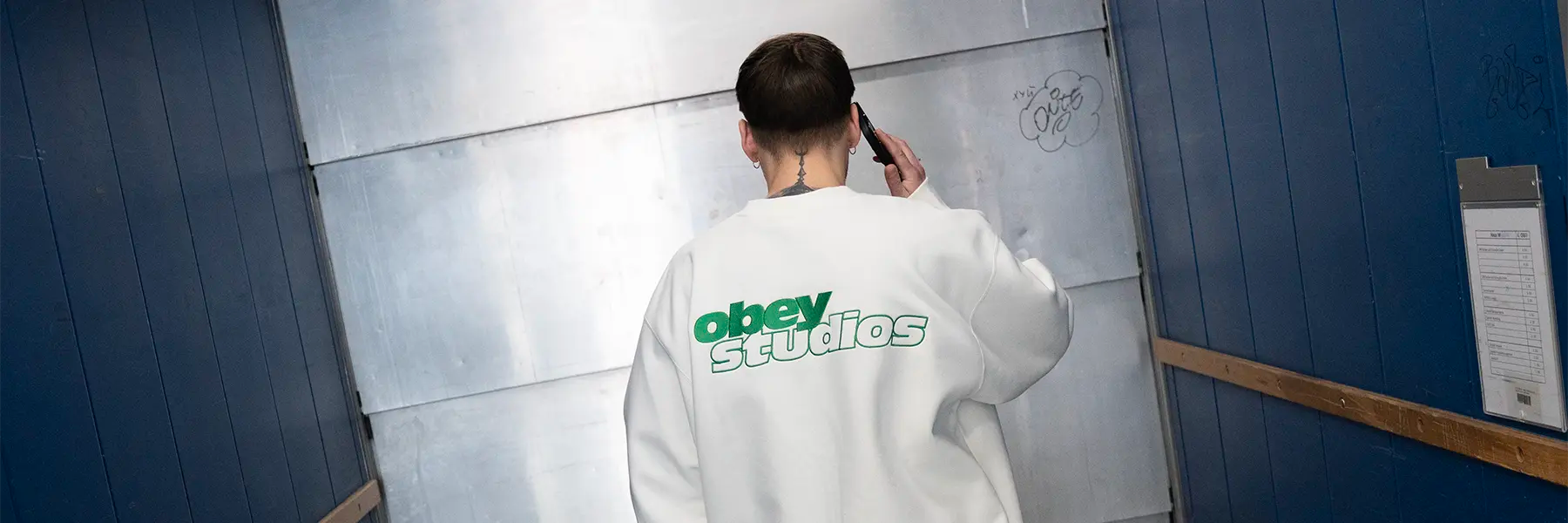 Person seen from behind wearing a white sweatshirt with 'obey studios' printed on the back walking in a corridor while holding a phone to their ear