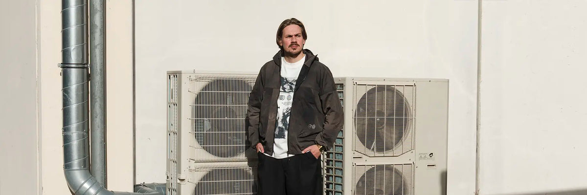 Man with mustache and beard wearing a black jacket from Purple Mountain Observatory and white graphic t-shirt standing with hands in pockets in front of two large air conditioning units