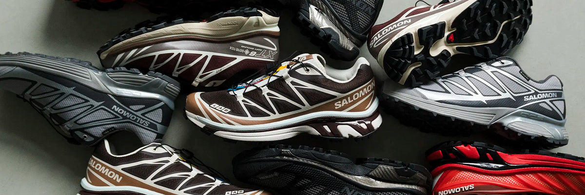 A group of Salomon trail running shoes in various colors and models, arranged closely together on a neutral gray surface. Visible models include the Salomon Speedo 600 in brown and white, the Enduro in gray and black, and a red and black model in the corner.