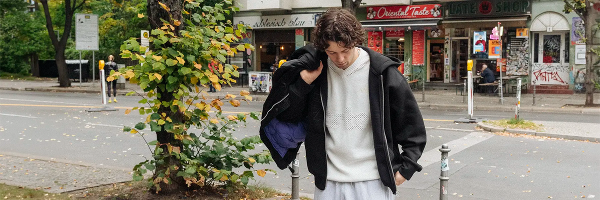 Man wearing a white perforated sweatshirt under a black zip-up jacket standing on a city sidewalk with autumn leaves