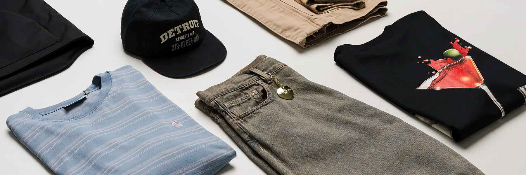 Casual Carhartt WIP clothing items arranged on a white surface, including a blue striped shirt, black cap, khaki pants, and a black T-shirt with a cocktail graphic.