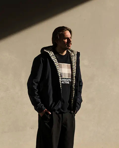 Man wearing a dark Aquascutum jacket with a checkered inner lining and a black shirt with a checkered pattern and text standing against a plain wall with hands in pockets