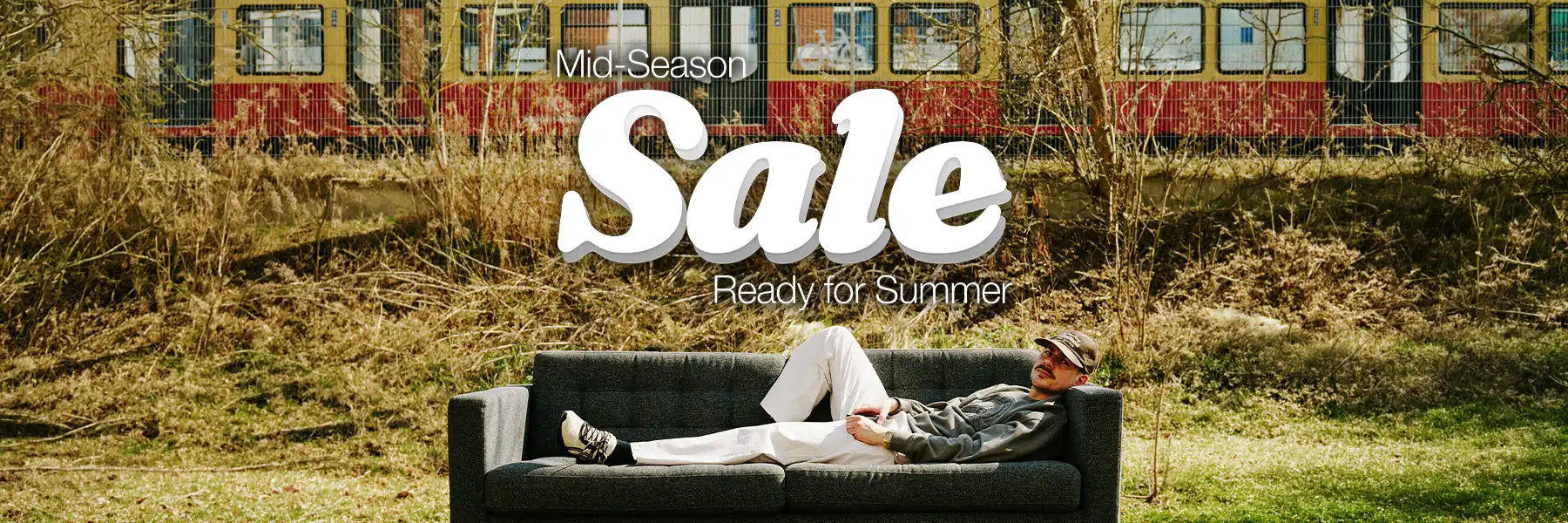 Man relaxing on a couch outdoors near a passing train. Text reads "Mid-Season Sale, Ready for Summer" in bold, stylish font, conveying a laid-back vibe.